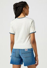 Camiseta blanca de manga corta con ribete azul, combinada con pantalones cortos de denim azul. Tejido liso, diseño ajustado, logo visible en el bolsillo trasero de los pantalones cortos.