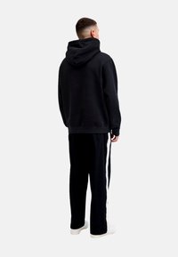 Schwarzer Kapuzenpullover aus strukturiertem Stoff, kombiniert mit schwarzen Hosen mit weißen Seitenstreifen. Modell trägt weiße Sneaker.