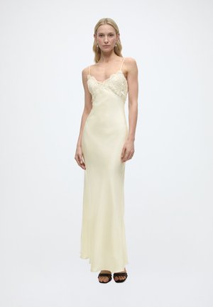 Γυναίκα που φοράει κρεμ σατέν φόρεμα με λεπτομέρειες δαντέλας στο μπούστο και λεπτόιιιιιιιιιιιιιιιι ς χονδροί ιιιιι ώμοιιιιιιιιιιιιιιιιι, συνδυασμένο με μαύρα πέδιλα με ανοιχτά δάχτυλα.