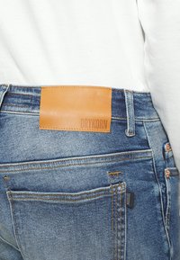 Bleka blå denimjeans med en beige läderlapp märkt "DRYKORN" vid midjan, med orange sömmar och en klassisk femficksdesign.