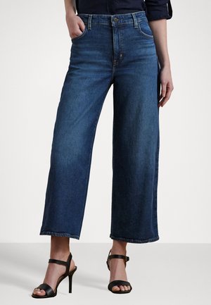 Vrouw draagt donkerblauwe wijde cropped jeans met zwarte hoge hakken sandalen, één hand in de zak, tegen een effen lichte achtergrond.