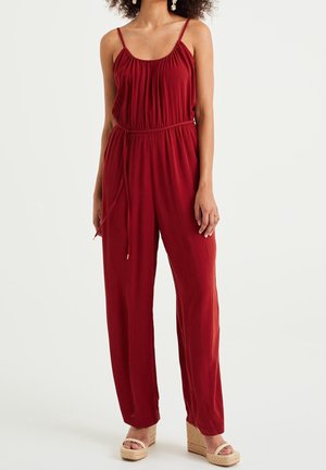 Kvinde iført rød ærmeløs jumpsuit med tynde stropper og kilehælssandaler, stående mod ensfarvet baggrund.
