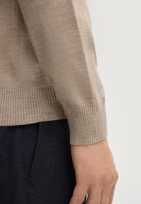 Beige Strickpullover mit gerippten Bündchen und Saum, der eine leicht strukturierte Oberfläche aufweist. Kombiniert mit enger geschnittenen, dunklen Stoffhosen.