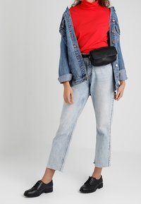 Žena v červeném roláku, s oversized džínovou bundou, světle modrými džíny, černou ledvinkou a černými oxfordkami stojící na jednobarevném pozadí.