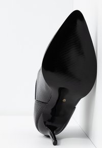 Sort højhælet sko med tekstureret overdel, der ligner reptilskind, og en blank sort sål. Hælen er tynd og spids med et gyldent tal "5."