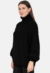 Młoda kobieta o długich włosach, ubrana w czarny golf z bufkami na rękawach, stojąca na tle jednolitego jasnego tła.