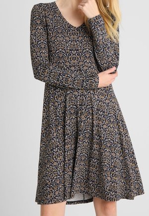 Femme portant une robe manches longues, longueur genou, avec un motif floral et de vigne sombre et complexe, posant avec une main sous le menton.