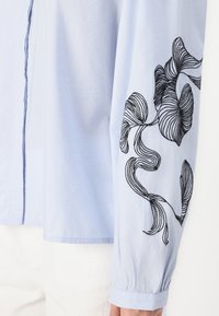 Blusa azul clara com mangas longas decoradas com padrão de folhas bordadas em preto. Tecido suave com fecho de botão na frente e punhos franzidos.