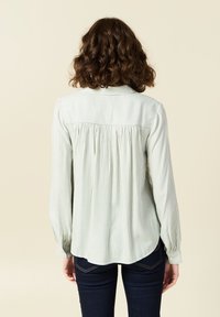 Hellgrün und weiß gestreiftes Langarmshirt mit Rückenfalten, abgerundetem Saum und Knopfverschluss am Hals. Glatte Stoffstruktur.