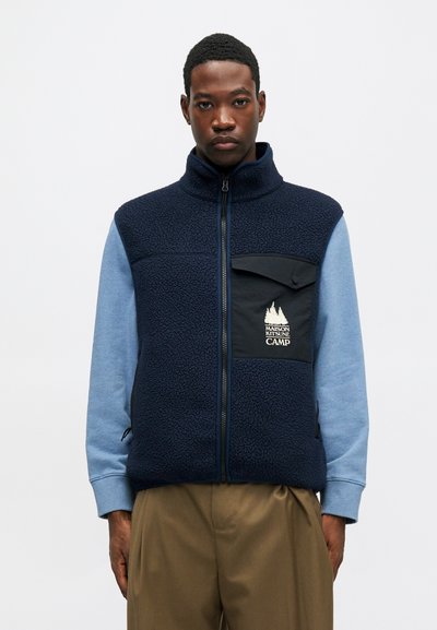 Námornícky fleecový vest s vysokým golierom, predným zipsom a čiernou kapsou so značkou. Modré rukávy a textúrovaný povrch.