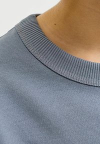 Κοντινό πλάνο σε γιακά πουλόβερ με στρογγυλή λαιμόκοψη από πλεκτό ύφασμα ανοιχτού γαλάζ ιου χρώματος και ένα μέρος δέρματος με μια μικρή ελιά κοντά στη γραμμή του λαιμού.