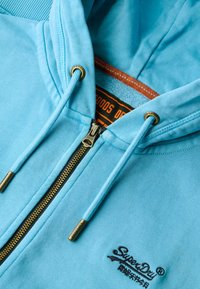 Sudadera con cremallera en azul claro, hecha de tela suave, que cuenta con una cremallera dorada, cordones con puntas de metal y un logo negro bordado en el frente.