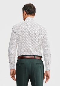 Biała koszula w czarne kropki, standardowy kołnierzyk, długie rękawy, zestawiona z zielonymi spodniami i brązowym paskiem widocznym z tyłu.