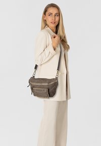 Sac bandoulière en cuir marron avec une surface texturée, une fermeture éclair sur le dessus, une poche avant et des clous décoratifs. Sangle ajustable pour une longueur personnalisable.