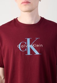 Burgundski pamučni majica s istaknutim svijetloplavim "CK" logom i tekstom "Calvin Klein" u bijeloj boji, s klasičnim okruglim izrezom.