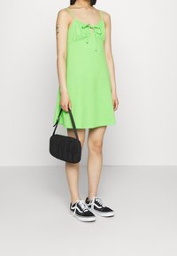 Grön, strukturerad klänning utan ärmar med detalj för knytning fram. Kombinerad med en svart, rektangulär handväska och svarta sneakers med vita sulor.