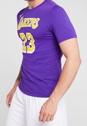 Mann trägt ein eng anliegendes lila Los Angeles Lakers-Trikot mit der Nummer 23 und weiße Shorts, steht vor einem einfachen grauen Hintergrund.