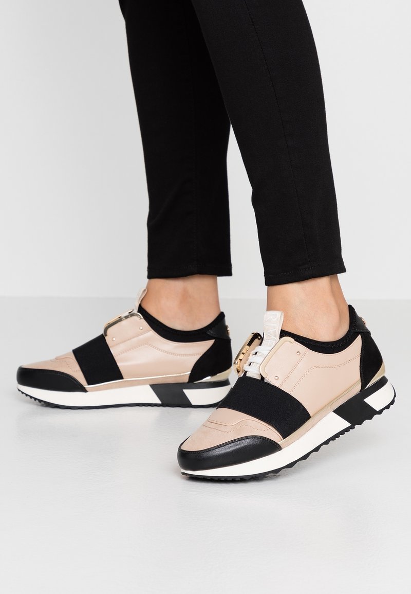 Baskets beiges et noires avec des bandes élastiques, accents en matériel doré, panneaux texturés et une semelle intermédiaire blanche. Présente un design sportif et une semelle en caoutchouc.