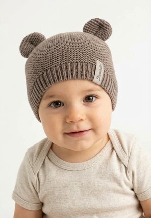Toddler with brown eyes wearing a knitted beige hat with round bear ears and a light beige short-sleeve shirt, smiling softly.