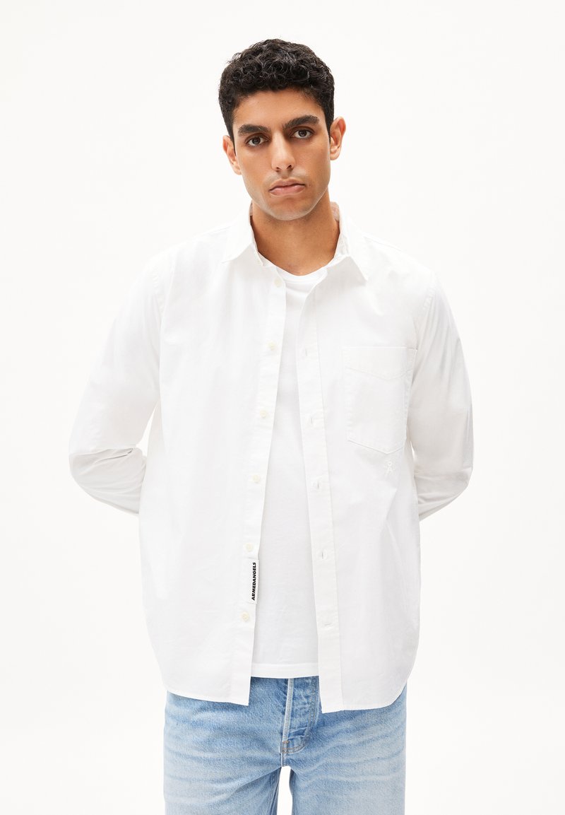 Camicia bianca a colletto, realizzata in cotone, con taschino sul petto e chiusura a bottoni, indossata sopra una maglietta bianca, abbinata a jeans in denim blu chiaro.