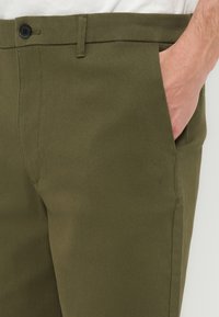 Κοντινό πλάνο ελιάς-πράσινου παντελονιού με ένα χέρι που έχει τοποθετηθεί μέσα στην μπροστινή δεξιά τσέπη, εμφανίζοντας λεπτομέρειες κουμπιού και θηλιάς για ζώνη.