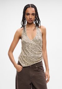 Χρυσό ασημί μπλουζάκι με ημίκλειστη λαιμόκοψη και ριχτό ύφασμα. Το μοντέλο το φοράει με καφέ παντελόνι, με τα χέρια στις τσέπες, σε ουδέτερο φόντο.