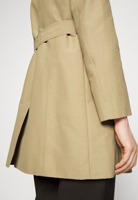 Beżowy płaszcz trench z dopasowanym krojem, z paskiem w talii i bocznymi rozcięciami. Wykonany z gładkiego materiału, z widocznymi szwami i długimi rękawami.