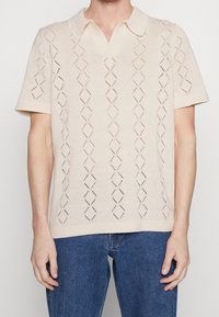 Stickad polo-shirt i ljusbeige med korta ärmar. Har ett diamantsmönster skapat av hålmönster och en klassisk krage.