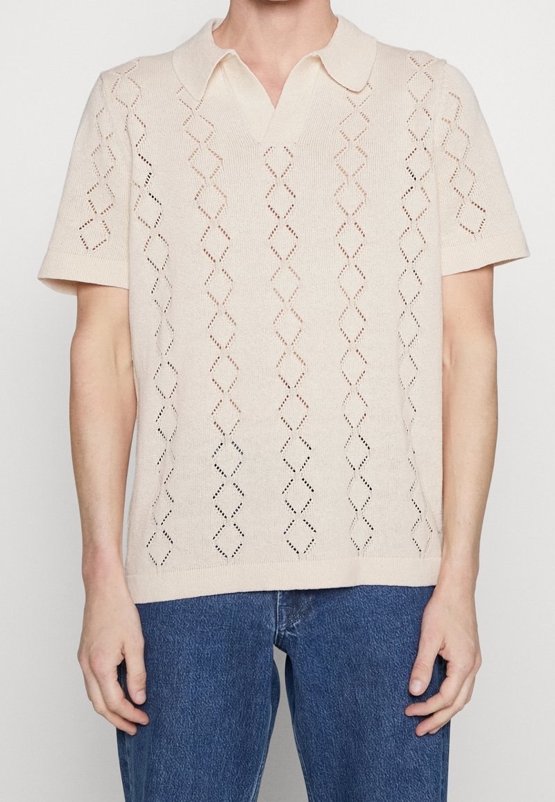 Stickad polo-shirt i ljusbeige med korta ärmar. Har ett diamantsmönster skapat av hålmönster och en klassisk krage.
