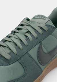 Green and gray sneakers with a textured fabric upper, flat laces, and a brown rubber sole. Features a prominent gray Nike logo.