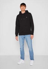 Felpa nera con cappuccio e tasca frontale, abbinata a jeans di denim azzurro chiaro e sneaker bianche, su uno sfondo grigio neutro.