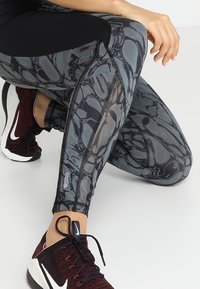 Osoba v kleče, oblečená v legínách s černobílým vzorem a tmavě červených teniskách Nike se bílým swoosh logem na bílém pozadí.