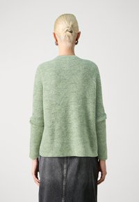 Osoba z blond włosami upiętymi w kok, nosząca srebrne kolczyki w kształcie kół, jasnoszary sweter o fakturze oraz ciemnoszarną spódnicę, stojąca tyłem do aparatu.