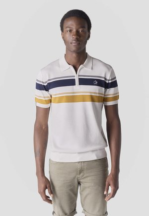 Młody mężczyzna stojący, ubrany w białą koszulkę polo z granatowo-musztardowymi paskami oraz beżowe, podwinięte spodnie, na tle jednolitego, jasnoszarego tła.