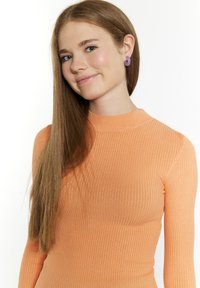 Lange mouwen, geribbelde top in lichtoranje, met een hoge hals. Model draagt paarse ronde oorbellen; haar is lang en steil.