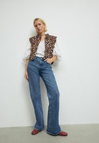 Luipaardprint vest, witte oogjesblouse, blauwe flared jeans en rode muiltjes met studs. Focus op gedurfde patronen en contrasterende materialen.