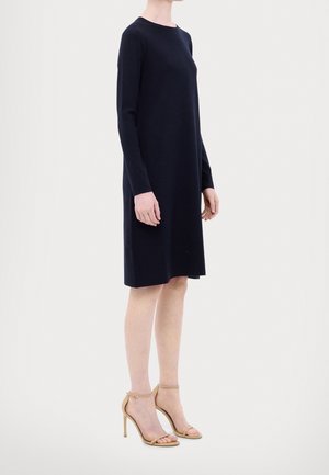 Femme portant une robe noire mi-longue à manches longues, associée à des sandales à talons hauts couleur nude, debout devant un fond clair uni.