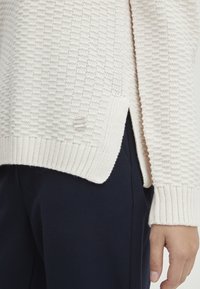 Pull en tricot crème avec un motif texturé, des poignets côtelés et des fentes sur les côtés. Présente un petit bouton logo transparent sur le côté gauche.