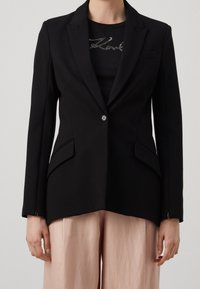 Blazer negro a medida con cierre de un solo botón, solapas marcadas, dos bolsillos delanteros y una silueta entallada, combinado con una camisa negra ajustada.