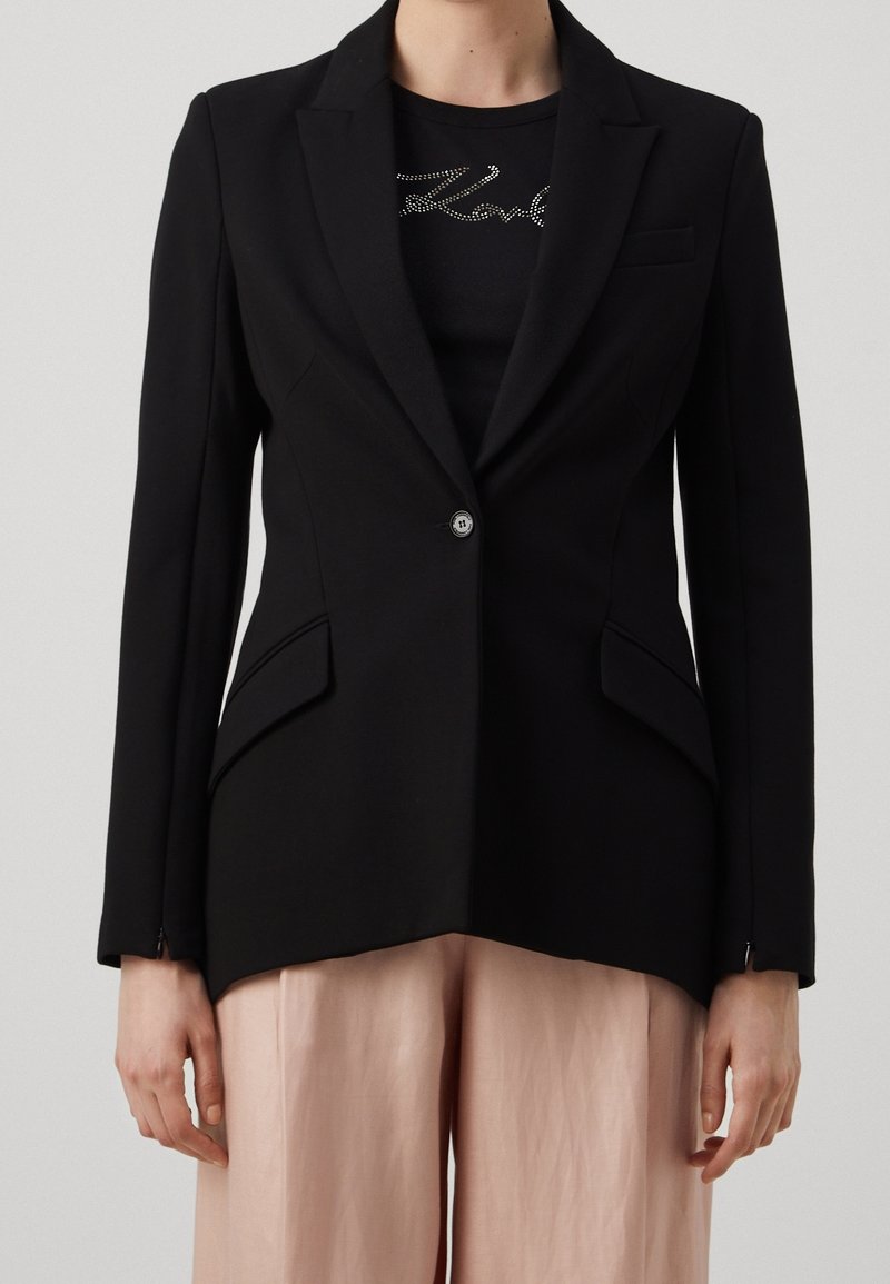 Blazer negro a medida con cierre de un solo botón, solapas marcadas, dos bolsillos delanteros y una silueta entallada, combinado con una camisa negra ajustada.