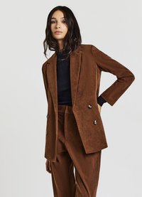 Blazer in corduroy marrone con doppio petto, bottoni dorati e rever a lancia, abbinato a pantaloni in corduroy coordinati e top nero a coste.