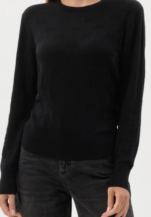 Modèle portant un pull noir à col rond avec un discret motif de texte et un jean gris foncé sur un fond blanc.