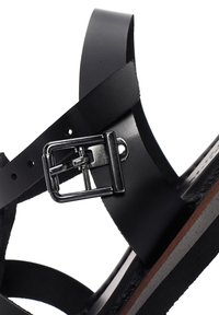 Black leather sandal featuring wide straps with a silver metal buckle, flat sole, and contrasting brown accents along the edge.