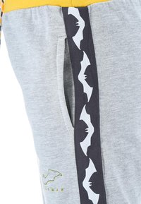 Graue Sweatpants mit gelbem elastischem Bund, Seitentaschen und schwarzem Seitenstreifen, der mit einem weißen Fledermaus-Logo-Muster verziert ist. Weicher Stoff.
