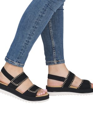 Füße in schwarzen Sandalen mit genähten Riemen und weißen Plateausohlen, kombiniert mit enganliegenden blauen Jeans.