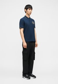 Άνδρας που στέκεται φορώντας μπλε σκούρο πόλο πουκάμισο με έμβλημα, μαύρο παντελόνι τύπου cargo και μαύρα αθλητικά παπούτσια, μπροστά σε απλό ανοιχτό φόντο.