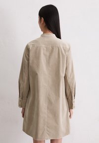 Frau mit langen, dunklen Haaren, die ein beiges, langärmliges, kragenbehaftetes Hemdkleid trägt und einer weißen strukturierten Wand gegenübersteht, mit entspannten Händen an den Seiten.