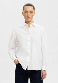 Camisa blanca de botones con cuello puntiagudo, mangas largas y tejido suave. Presenta pequeños botones de color claro y un corte relajado.