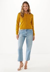 Žena se usmívá, má na sobě hořčicově žlutý cardigan na knoflíky, světle modré džíny s vysokým pasem a zlaté otevřené lodičky, stojí na bílém pozadí.