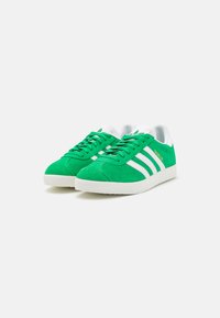 Green suede sneakers with white stripes, flat laces, and a textured rubber sole. Features a padded collar and "Gazelle" branding on the side.